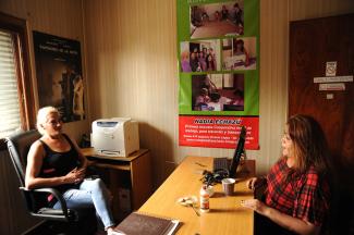Brisa Escobar sitting behind her desk, in conversation with a woman. The visitor has long brown hair and is wearing a top with red and black pattern