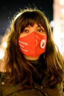 The photo shows Sopo Japaridze, one of the co-founders of the Solidarity Network Union. Sopo has long brown hair, with bangs, and brown eyes, and wears a red mask of the Solidarity Network Union. The picture is taken at night.